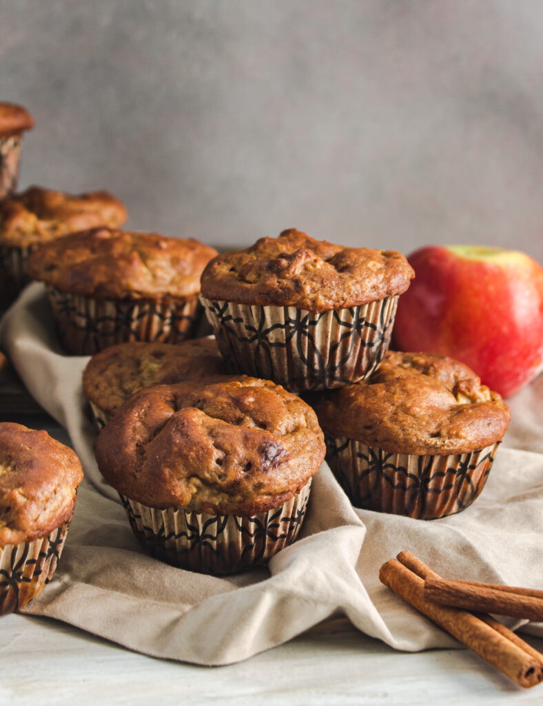Pile of muffins stacked on a beige napkin with cinnamon sticks in front and a whole apple behind