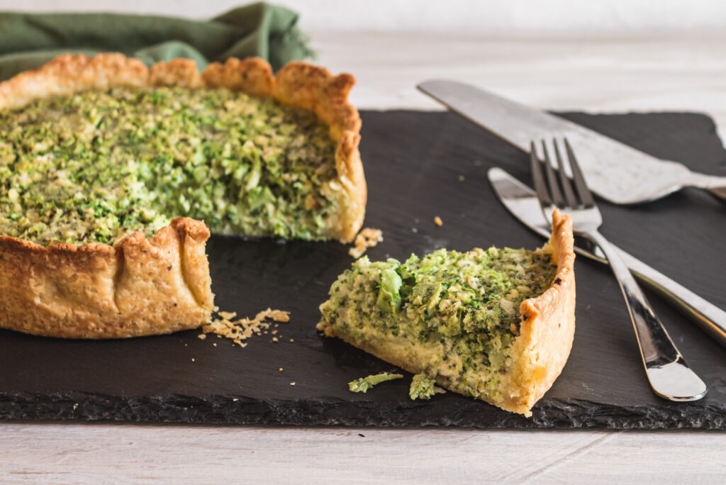 Quiche with a slice cut out on a rectangular slate board with a knife, fork, and cake server to the side.