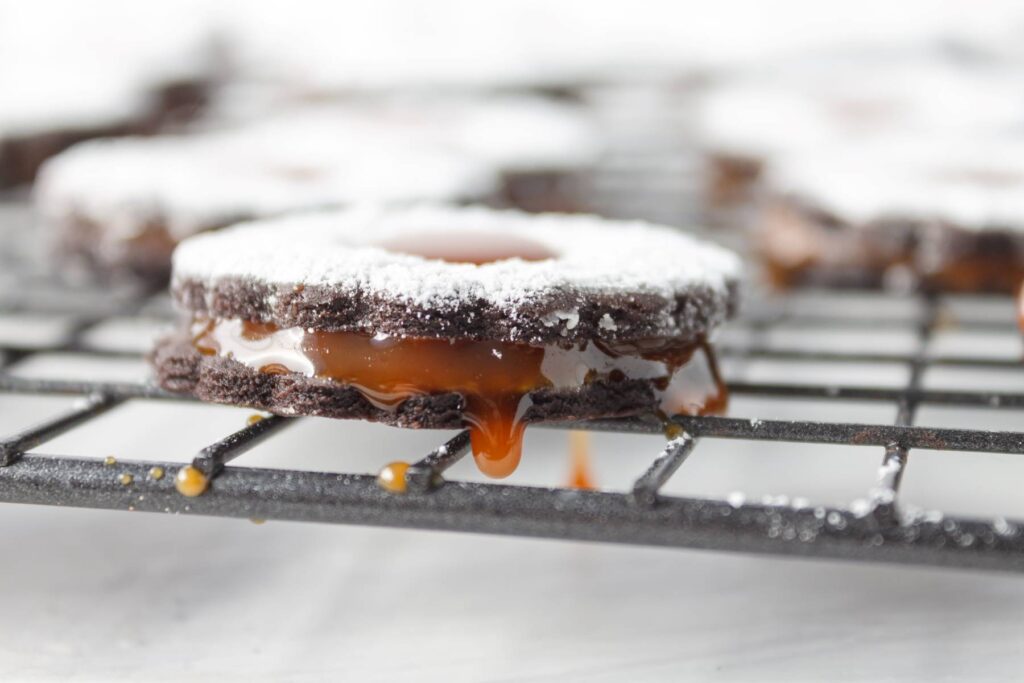 Chocolate and caramel sandwich cookies dusted with powdered sugar sitting on cooling rack and dripping caramel.