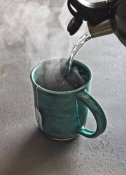 Hot water pouring from kettle into tourquise mug with steam billowing out.