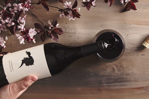 Top view of a hand pouring a bottle of red wine into a glass on a table with branches of plum blossoms around