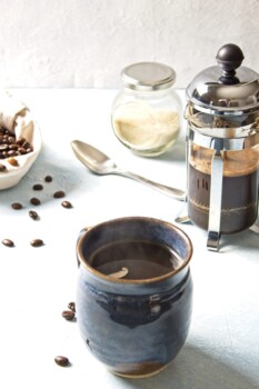 Blue mug with slight steam coming out of top in front of a small french press and sack of coffee beans. On a light blue background with scatter coffee beans