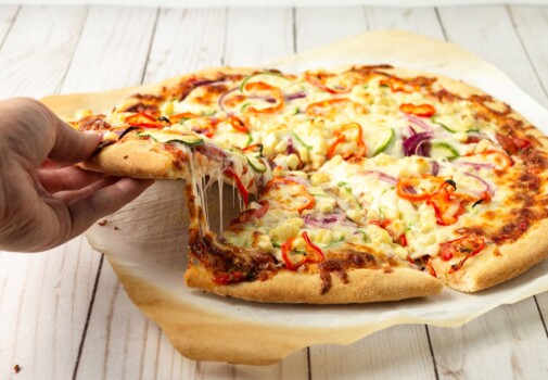 White hand lifting a slice of vegetable pizza away from the whole pizza resting on parchment paper